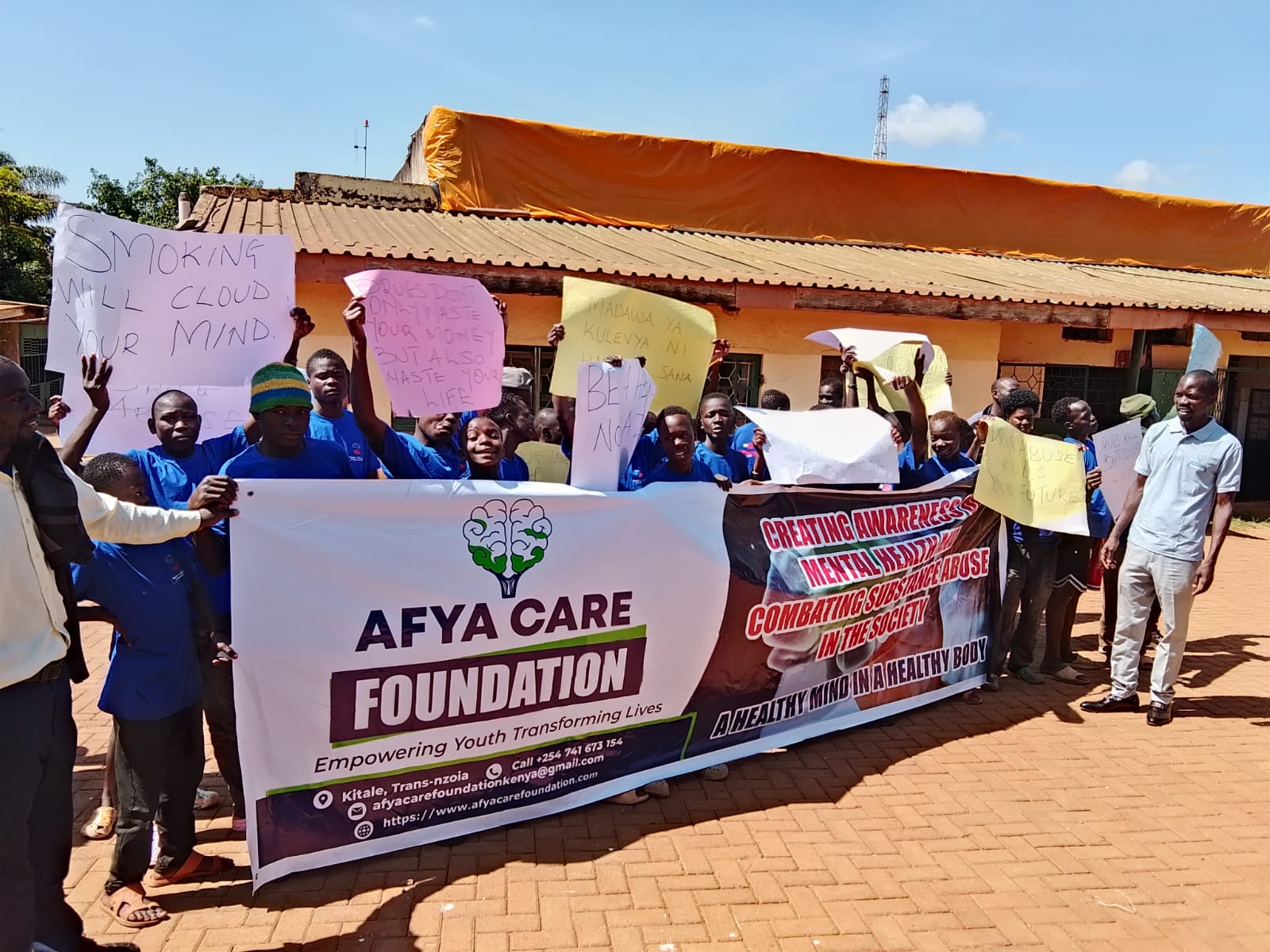 Young people and Afya Care Foundation staff behind a foundation banner reading Empowering Youth Transforming Lives, holding handwritten placards in English and Swahili about the dangers of smoking and drug abuse